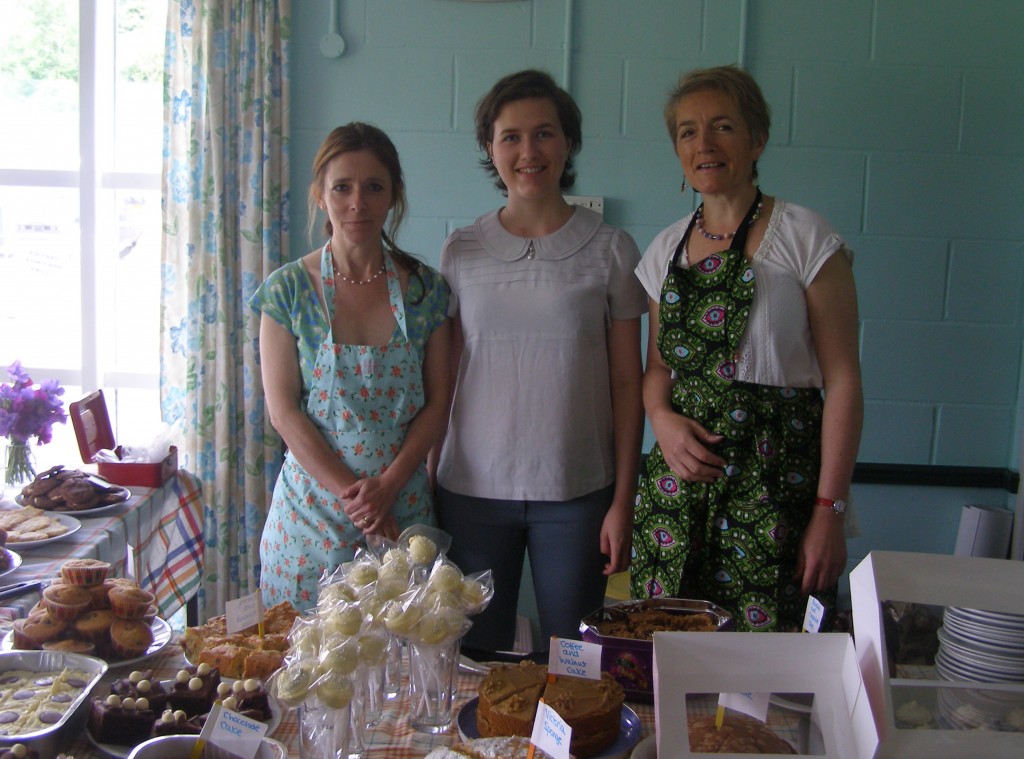Three ladies organising a cake sale, stand in front of table full of cakes.